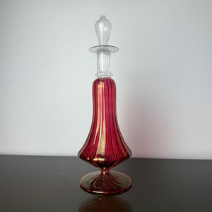 May include: A decorative glass perfume bottle with a clear stopper. The bottle is a deep red color with vertical gold stripes. The base and stopper are clear glass.