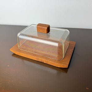 Puede incluir: Una mantequera rectangular con tapa transparente en forma de cúpula y base de madera. La tapa tiene un pequeño tirador rectangular de madera. La base también es de madera, con forma ligeramente curvada.