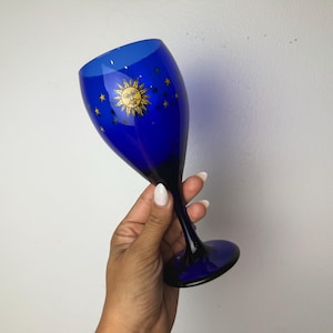 May include: A cobalt blue glass goblet featuring a gold-coloured sun face graphic surrounded by small gold stars. The stem and base are also made of cobalt blue glass, creating a uniform colour throughout the entire piece of drinkware.