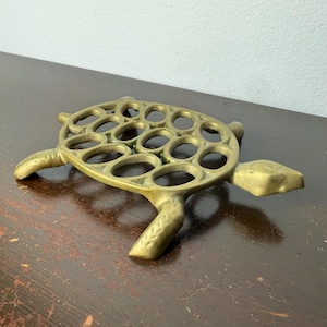 May include: A brass turtle-shaped trivet with circular cutouts on its shell. The trivet has four legs and a head and tail, all in a matching brass finish. It is designed to protect surfaces from hot cookware, a decorative kitchen item.
