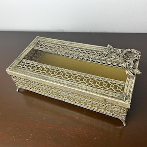 Puede incluir: Un porta pañuelos de metal plateado con un diseño floral decorativo. El porta pañuelos tiene un diseño de malla y una forma rectangular.