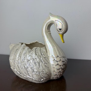May include: A ceramic swan planter featuring an iridescent, off-white glaze with a glossy finish. The swan has a long, curved neck, a bright yellow beak, and detailed molded feather textures covering its body. The top of the swan is open.