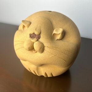 Könnte beinhalten: Eine tanfarbene Keramik-Katzenfigur mit einem braunen Schnurrbart und geschlossenen Augen. Die Katze hat einen runden Körper und eine strukturierte Oberfläche.