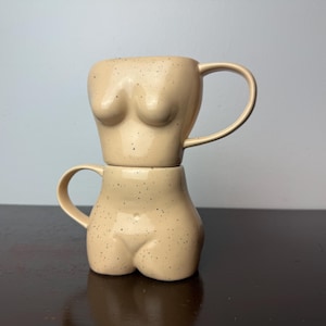 May include: Two beige ceramic mugs stacked, each shaped like a torso. The top mug is the upper body, and the bottom mug is the lower body. Both mugs have a speckled finish and a curved handle.