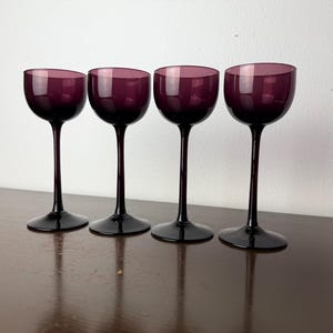 May include: Four stemmed wine glasses with a deep purple bowl and a black stem and base. The glasses are arranged in a row on a dark brown surface, reflecting the light. The background is a plain white wall.