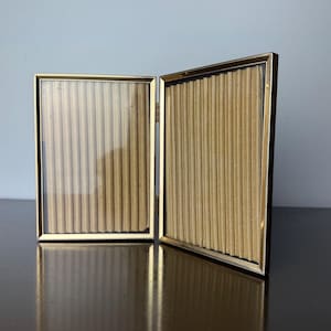 Op de afbeelding: Een goudkleurig, scharnierend dubbel fotolijstje. Het frame is open en onthult twee rechthoekige secties met een getextureerde, verticaal gestreepte achterkant. Het frame heeft een dunne, gouden rand en een donkere achterkant. Het frame wordt op een donkere ondergrond weergegeven.