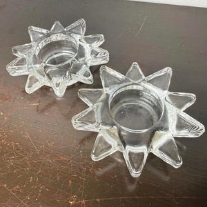 May include: Two clear glass candle holders shaped like multi-pointed stars. Each holder features a circular center depression designed to hold a candle, surrounded by radiating, faceted points that create a distinct sunburst silhouette.
