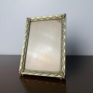 May include: A vintage rectangular picture frame with an ornate, scalloped edge design. The frame is made of a gold-colored metal and has a simple, neutral backing. It stands on small feet and is angled slightly.
