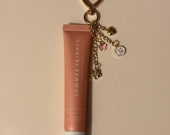 Puede incluir: Un tubo rosa de bálsamo labial Summer Fridays con una cadena dorada y abalorios. Los abalorios incluyen un corazón, una mariposa y una letra N.