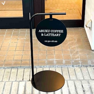 Puede incluir: Letrero exterior de metal negro para una cafetería. El letrero tiene un panel circular negro con el texto "ABUKU COFFEE & LATTEART" y el horario de apertura. El letrero está sostenido por un poste curvo y una base redonda.