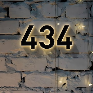 May include: Black illuminated house number 434 against a white brick wall. The numbers are large and modern, with a warm, yellow glow outlining them, creating a striking contrast against the textured brick.