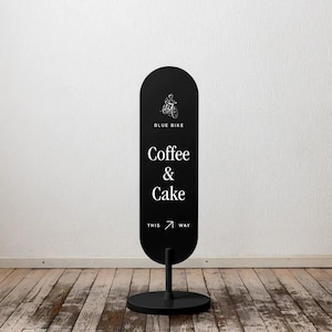 May include: Black sign with the text "BLUE BIKE Coffee & Cake" and an illustration of a cyclist. The sign is mounted on a black stand and is pointing the way.