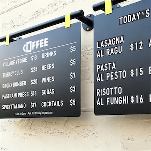 May include: Two black rectangular menu boards with white text and prices, suspended from a black metal bar with yellow clips. The left board lists coffee, sandwiches, and drinks with prices. The right board displays daily specials like lasagna, pasta, and risotto.