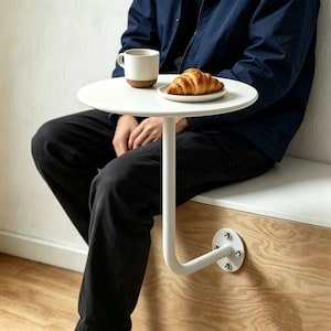 Puede incluir: Una mesa auxiliar redonda blanca con un brazo de soporte de metal blanco curvado. La mesa está fijada a una superficie de madera con una placa de metal y pernos. Una taza de café y un cruasán en un plato pequeño están sobre la mesa.