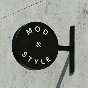May include: A black, circular sign with the words "MOD & STYLE" in white lettering. The sign is mounted on a black metal bracket against a gray concrete wall. The sign is a shop sign.