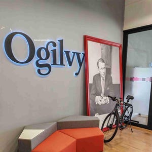 Puede incluir: Una foto en blanco y negro de un hombre con traje y corbata está enmarcada en un marco rojo. La foto está colgada en una pared con un letrero negro que dice "Ogilvy" con luces azules alrededor de las letras. Un sofá gris y naranja está en primer plano con una bicicleta negra apoyada en la pared.