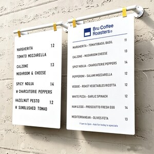 May include: Two white rectangular menu boards suspended from white horizontal bars with yellow clips. The boards feature black text listing food items like Margherita, Calzone, and Spicy Nduja with prices, and the Bru Coffee Roasters logo at the top.