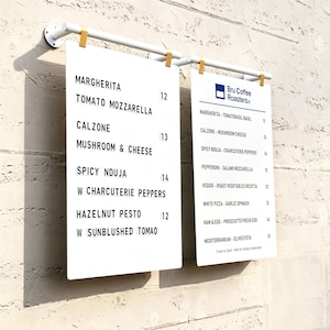 May include: Two white rectangular menu boards hanging from a white metal rod. The left board lists Margherita, Tomato Mozzarella, Calzone, Mushroom & Cheese, Spicy Nduja w Charcuterie Peppers, and Hazelnut Pesto w Sunblushed Tomao with prices.