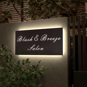 May include: A rectangular black sign with the words "Blush & Breeze Salon" in elegant white script. The sign is illuminated with a warm, soft light, and is mounted on a dark gray wall. Green plants are visible in the foreground.