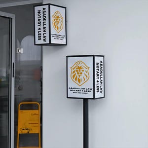May include: Two white and black rectangular signs with the text "ASADULLAH LAW NOTARY 4 LESS" in black. Each sign features a gold lion crest. One sign is mounted on a pole, the other is mounted on a wall.
