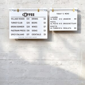 May include: Two white rectangular menu boards with black text hanging from white and gold metal bars. The left board lists coffee and food items like Village Veggie and Turkey Club with prices. The right board displays Today's Menu with Italian dishes.