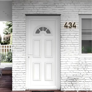 May include: A white front door with a half-circle window at the top and four rectangular panels. The number "434" is displayed on the brick wall. The door is set against a white brick wall, with a small window and a white chair visible.