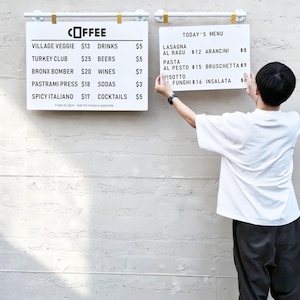 May include: Two white rectangular menu boards with black text mounted on a wall. The left board lists coffee, food items, and prices. The right board is titled TODAY'S MENU and lists various Italian dishes with prices in dollars.