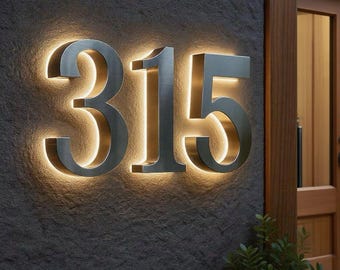 Puede incluir: Números de casa iluminados '315' en metal cepillado, montados en una pared de piedra gris oscuro. La retroiluminación cálida crea un aspecto moderno y elegante para cualquier hogar.