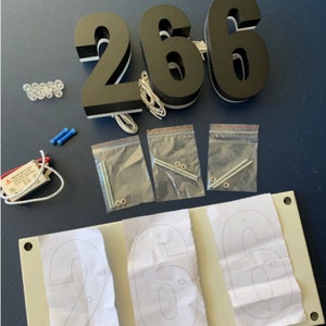 May include: Black illuminated house numbers, "266", with white trim and wiring. Includes mounting hardware, LED lights, and a transformer. A template with the numbers is also included.