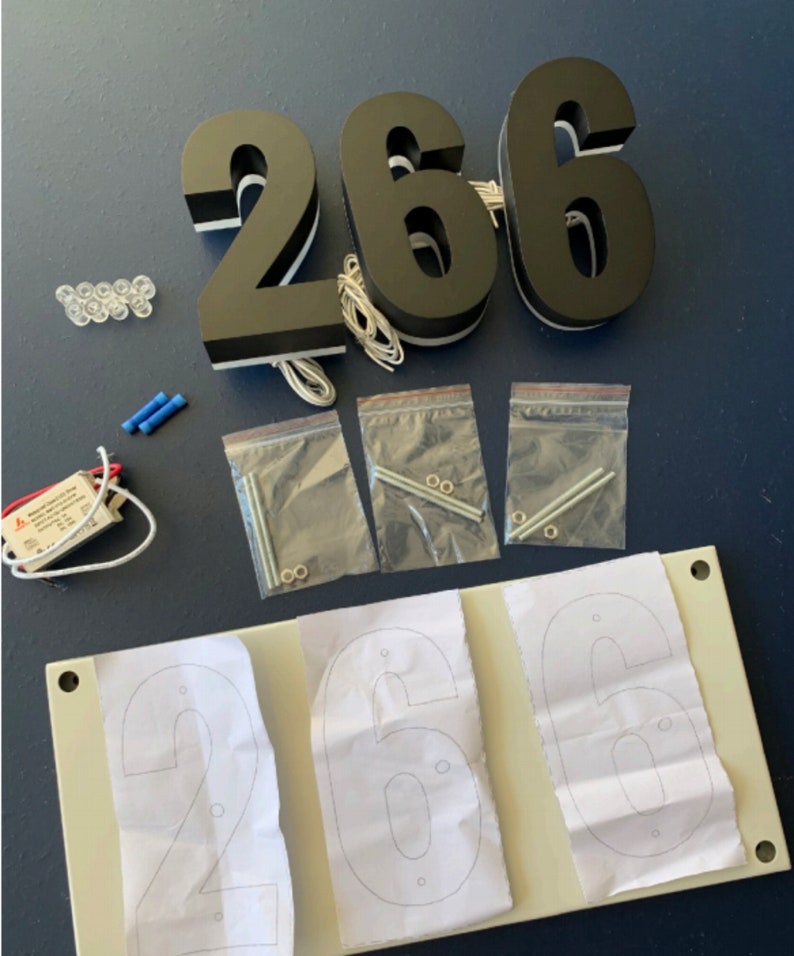 May include: Black illuminated house numbers, "266", with white trim and wiring. Includes mounting hardware, LED lights, and a transformer. A template with the numbers is also included.