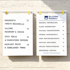 May include: Two white rectangular menu boards with black text hanging from white bars with yellow clips. The left board lists pizza options with prices. The right board features the Bru Coffee Roasters logo and a list of pizzas with prices.