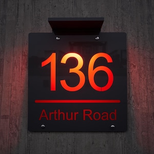 Puede incluir: Letrero de dirección negro con los números rojos iluminados "136" y el texto "Arthur Road". El letrero está montado en una pared de madera. Los números y el texto están en una fuente negrita y moderna. El letrero tiene un diseño elegante y contemporáneo.
