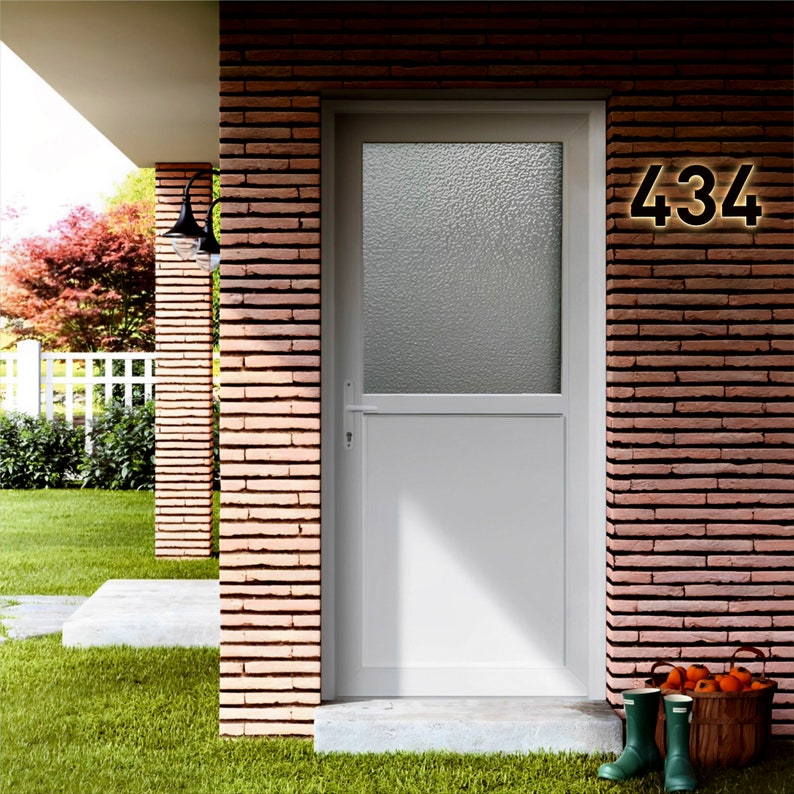 May include: A white exterior door with a textured glass panel and the number 434 illuminated in gold. The door is set in a brick wall. Green rubber boots and a basket of fruit sit on the doorstep. A black light fixture is mounted on the brick wall.