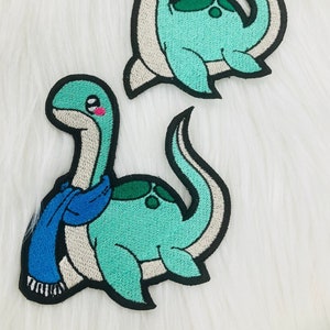 May include: Two embroidered iron-on patches of a cartoon plesiosaur. One plesiosaur is wearing a blue scarf. Both are green and white with black outlines.