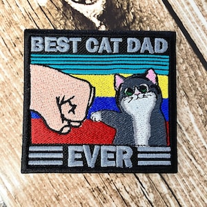 May include: Embroidered patch with a retro design featuring a fist bumping a cat. The text reads "Best Cat Dad Ever" in gray and white lettering.