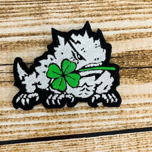May include: Embroidered patch depicting a white and black horned animal holding a green four-leaf clover. The patch has a black border and is set against a wooden background. Suitable for St. Patrick's Day.