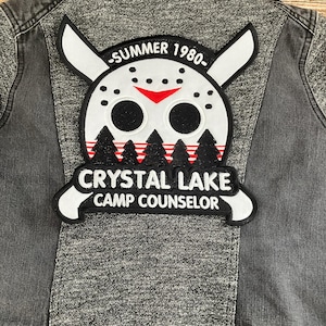 Peut inclure: Une veste en jean noire avec un écusson brodé blanc et rouge qui dit "Summer 1980" et "Crystal Lake Camp Counselor". L'écusson représente un masque de hockey avec une barre rouge et deux yeux noirs.
