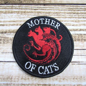 May include: A black and white embroidered patch with a red three-headed cat design. The text "Mother of Cats" is embroidered in white around the design.