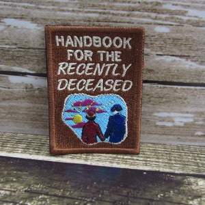May include: Brown embroidered patch with the text "Handbook for the Recently Deceased" and an illustration of two figures walking towards a sunset with clouds.