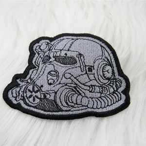 May include: A black and gray embroidered patch depicting a detailed illustration of a power armor helmet. The helmet has a visor, a large air filter, and a complex system of pipes and tubes.
