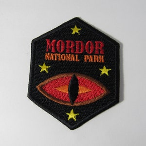 May include: A black hexagonal patch with the text "Mordor National Park" in red and orange lettering. A red eye with an orange pupil and a brown eyebrow is in the center of the patch. Five yellow stars surround the eye.