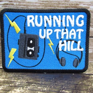 May include: Blue embroidered patch with a cassette tape, headphones, and lightning bolts. The text "RUNNING UP THAT HILL" is embroidered in white.