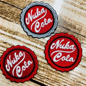 May include: Three circular embroidered patches shaped like bottle caps. Each patch features a vibrant red background with the white text Nuka Cola written in a stylized script font. The edges are scalloped, with one patch having a grey border.