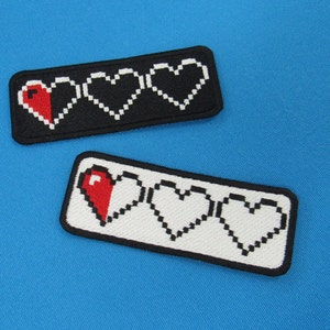 May include: Two embroidered patches depicting a pixelated heart health bar. The top patch has three hearts, one red and two black. The bottom patch has three hearts, one red and two white.