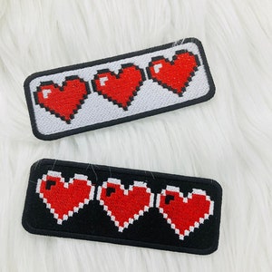 Peut inclure: Deux patchs brodés avec trois cœurs rouges pixelisés sur un fond blanc et noir. Les patchs sont rectangulaires et ont une bordure noire.