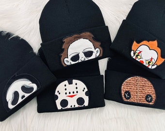 May include: A collection of five black knit beanies, each featuring a unique embroidered patch of a horror movie character. The patches depict a white ghost mask, a brown-haired figure, a hockey mask, a clown face, and a burlap sack mask.