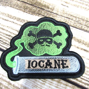 May include: Black and green embroidered patch with a skull and crossbones inside a green cloud and the word "IOCAINE" on a silver tube.