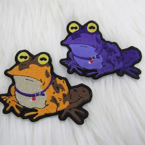 May include: Two embroidered frog patches. One is orange and brown, the other is purple. Both have yellow eyes and are wearing a collar with a small red heart. The patches are on a white, fluffy surface.