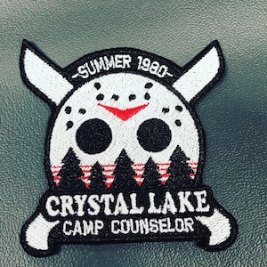 May include: White and black embroidered patch featuring a hockey mask, pine trees, and the text "Summer 1980", "Crystal Lake", and "Camp Counselor".