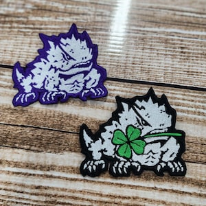 May include: Two embroidered patches featuring a stylized creature with sharp spikes. The top patch has a white creature on a purple background. The bottom patch features a white creature on a black background, holding a green four-leaf clover in its mouth.